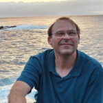 Johnson smiles as he sits outdoors on a boat on the water with the sea behind him.