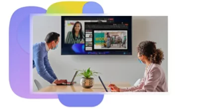Two people in a conference room wearing PPE while participating in a Teams video call being displayed on a large television.