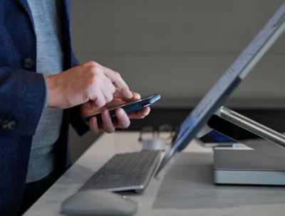 A person’s hands using a mobile phone in front of a desktop monitor.