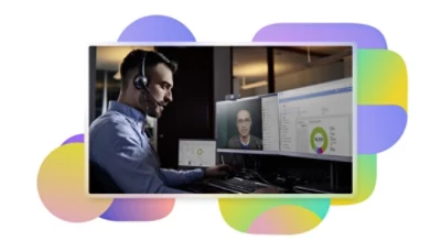 A person sitting at their desk wearing a headset participating in a Teams video call being displayed on one screen and looking at data on another.