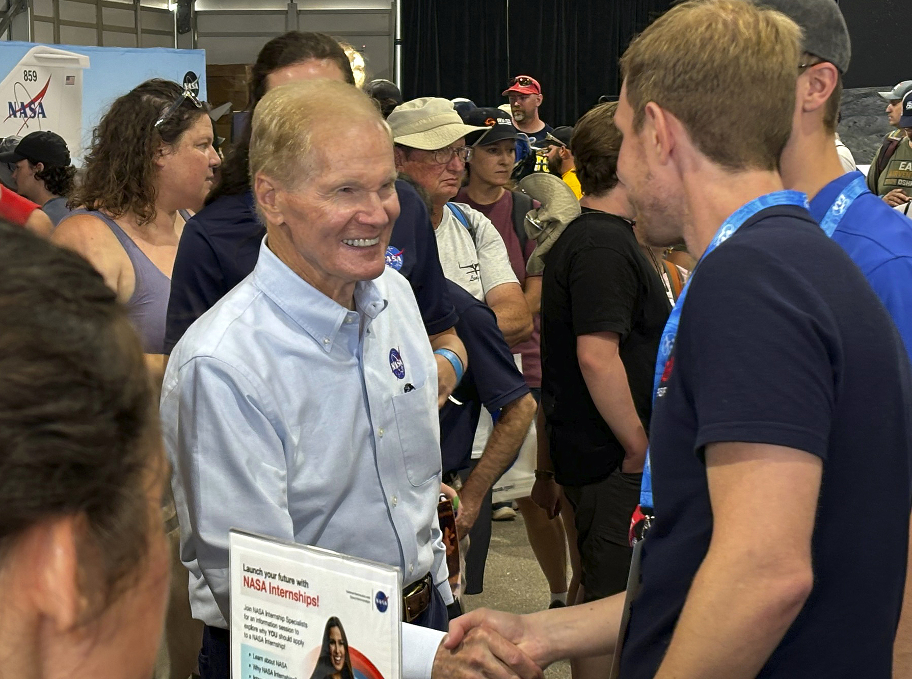 A man shake hands with another man as a group of people mill about behind them.