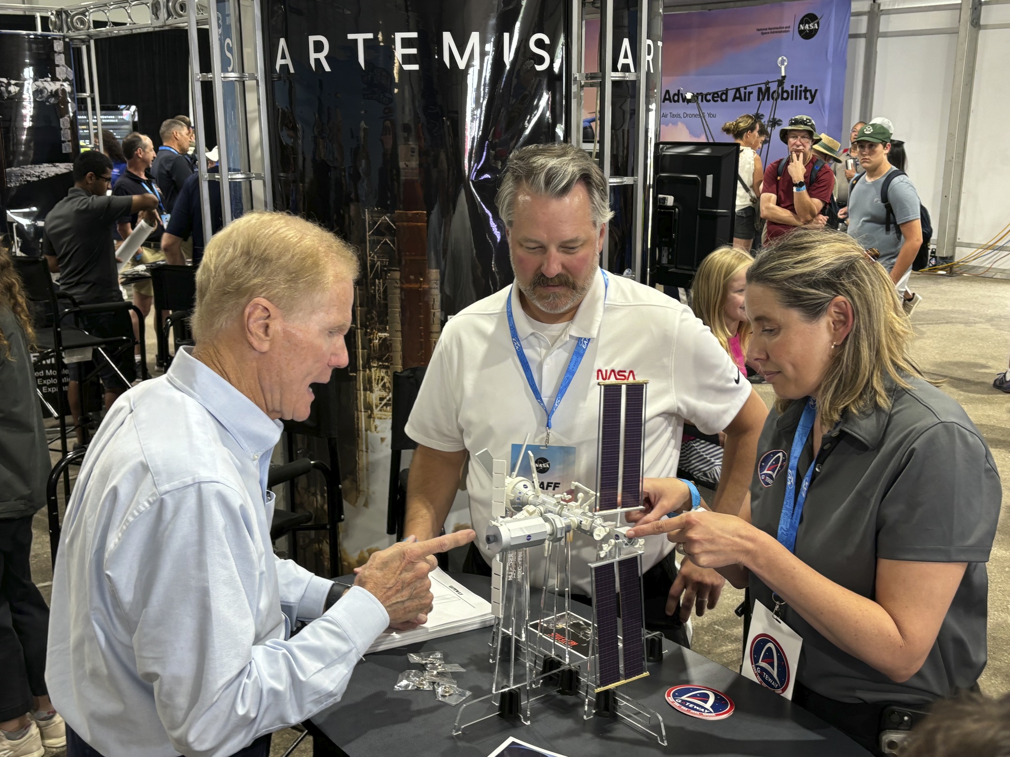 Three people stand around a table on which a model of a space station sits.
