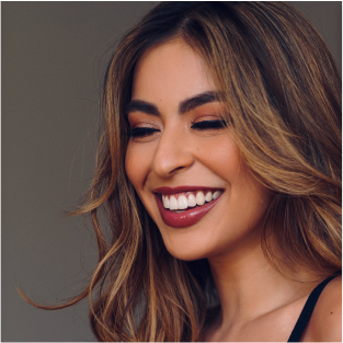 A woman with long hair is smiling with her eyes closed against a plain background.