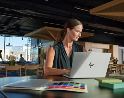 A woman with a ponytail sits at a table in a modern office space, working on an HP laptop
