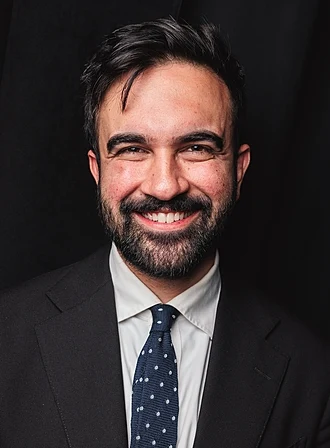 A picture of Zohran Mamdani. He is smiling and looking directly into the camera. He is wearing a black suit and a blue, white spotted tie.