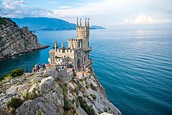 Vogelperspektive auf die Festung, die auf die Klippe gebaut wurde. Die Festung besteht aus hellem Stein und mehreren kleinen Türmen, die auf dem großen runden Turm und dem eckigen Gebäude verteilt sind. Viele Touristen haben sich auf der Plattform vor der Festung versammelt. Im Hintergrund sieht man das Meer und die Felsenküste.