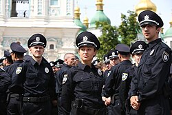 Im Vordergrund der Farbfotografie stehen drei Polizisten, jeweils ein Mann links und rechts vom Foto und eine Frau in der Mitte, mit Blick an der Kamera vorbei. Im Hintergrund sind weitere Polizisten und eine orthodoxe Kirche zu erkennen. Alle Polizisten tragen die schwarze Uniform mit einem runden Abzeichen an ihrer linken Brust.