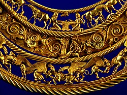 Ausschnittfoto einer goldenen Brustkette, die in drei waagerechten Szenen aufgeteilt ist, wobei der untere und obere Teil im Hintergrund durchsichtig sind. Somit erkennt man den dunkelblauen Untergrund. Im unteren Teil kämpfen geflügelte Wesen mit Pferden, der Mittelteil wird durch Blumen verziert und im oberen Teil sind Alltagsszenen mit Menschen und Pferde zu sehen.